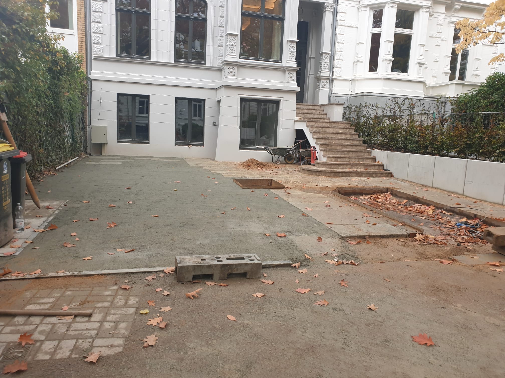 Przygotowanie terenu przed budynkiem z dekoracyjną fasadą. Częściowo wyrównany teren, widoczne narzędzia i liście. Etap prac ziemnych przed ułożeniem nawierzchni.