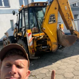 Żółta koparko-ładowarka JCB na placu, widziana z bliska w kadrze selfie z uśmiechniętym mężczyzną wskazującym palcem, budynek w tle.