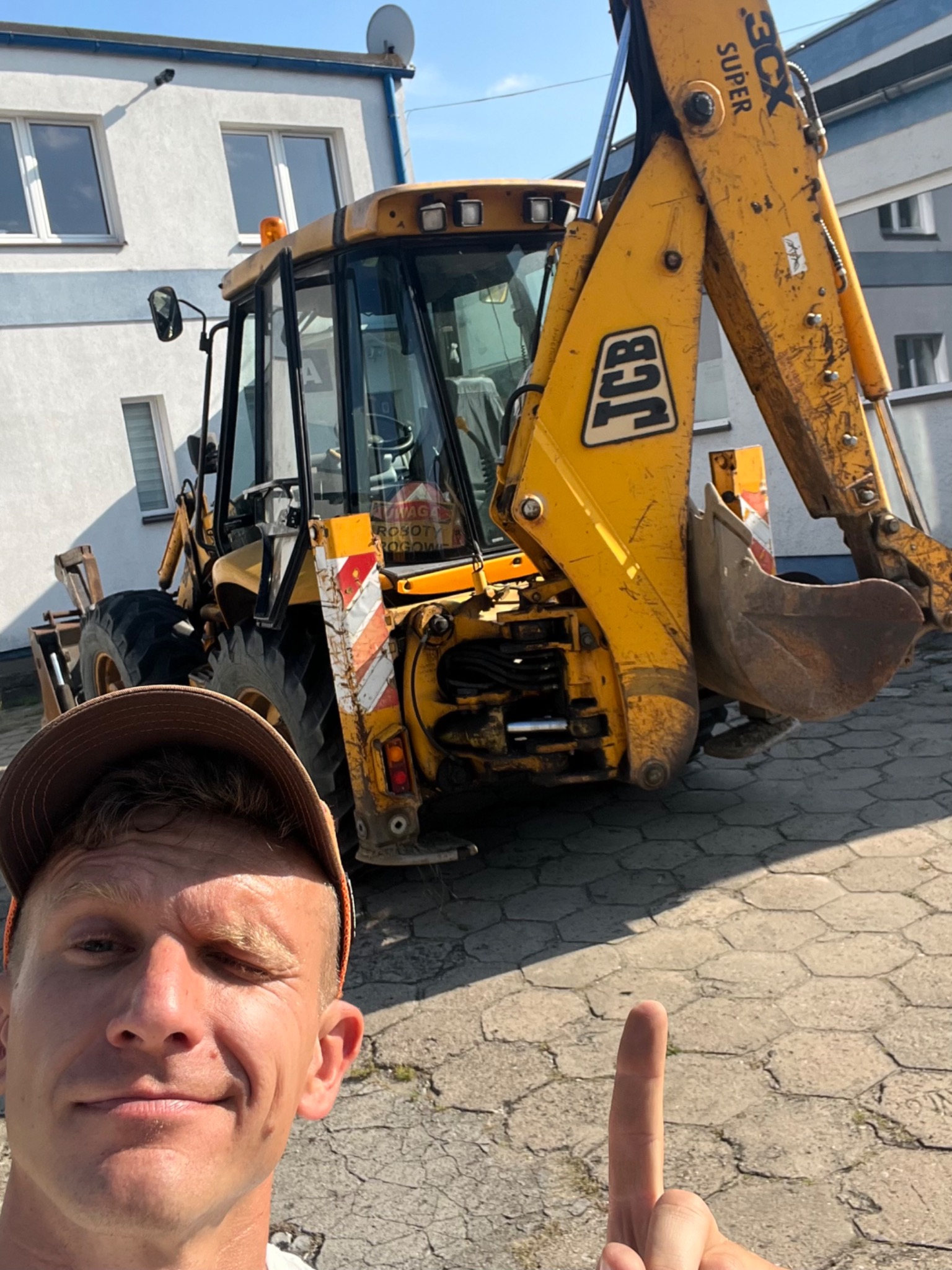 Żółta koparko-ładowarka JCB na placu, widziana z bliska w kadrze selfie z uśmiechniętym mężczyzną wskazującym palcem, budynek w tle.