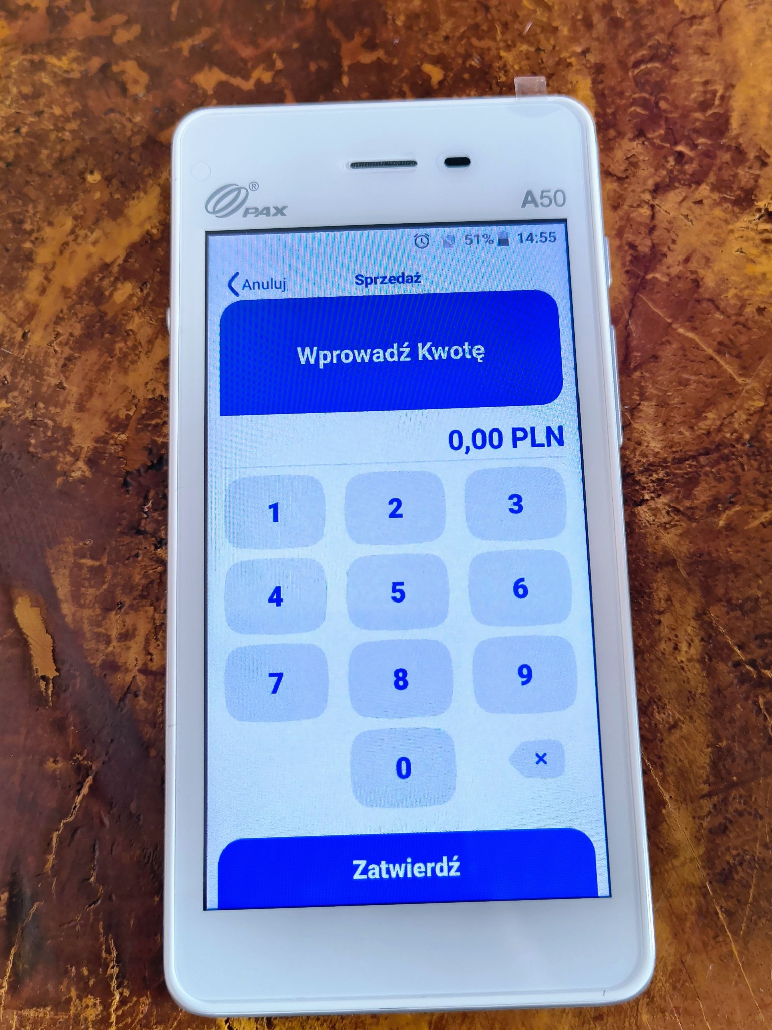 Terminal płatniczy PAX A50 z ekranem wprowadzania kwoty, widoczne logo PAX. Urządzenie leży na brązowym blacie z wyraźną fakturą.