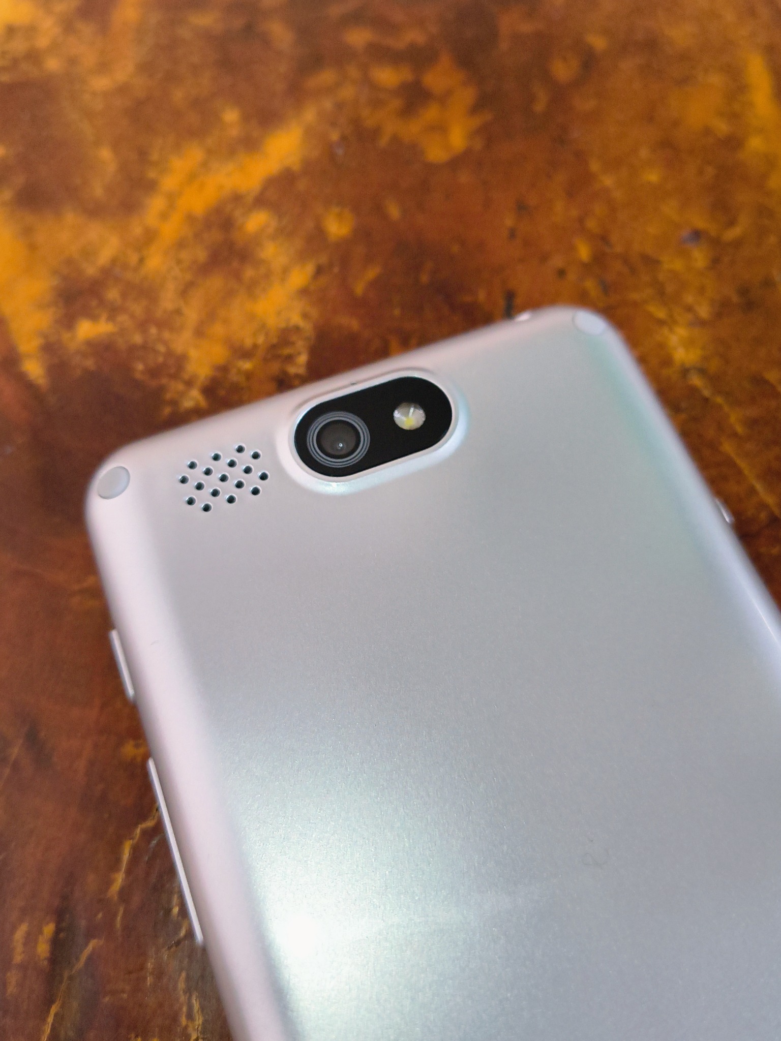 Zbliżenie na srebrny smartfon z widocznym aparatem i głośnikiem na tle abstrakcyjnej, ciemnej powierzchni z żółtymi wzorami.