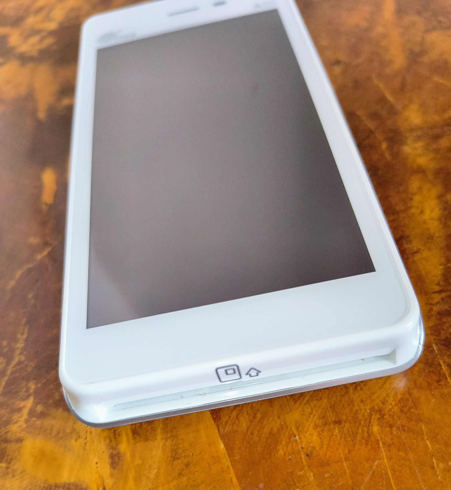 Biały smartfon leżący na drewnianym stole, zbliżenie na dolną część obudowy z widocznymi ikonami, ekran wygaszony, widoczna faktura drewna.