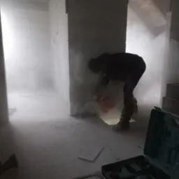 Praca w trakcie: robotnik tnie bloczki piłą elektryczną w zapylonym, surowym wnętrzu. Widoczny kufer z narzędziami i poziomica.