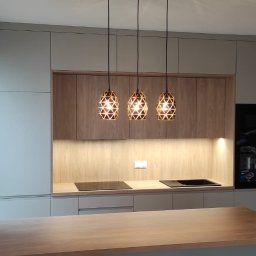 Matheo wood meble - Nowoczesna kuchnia z drewnianymi frontami i trzema designerskimi lampami. Jasne szafki, płyta indukcyjna i piekarnik w zabudowie. Widoczna wyspa kuchenna.