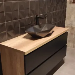 Nowoczesna szafka łazienkowa z umywalką w kształcie rombu i czarną armaturą na tle ściany z geometrycznych płytek. Fronty szafki w kolorze czarnym, blat w odcieniu drewna.