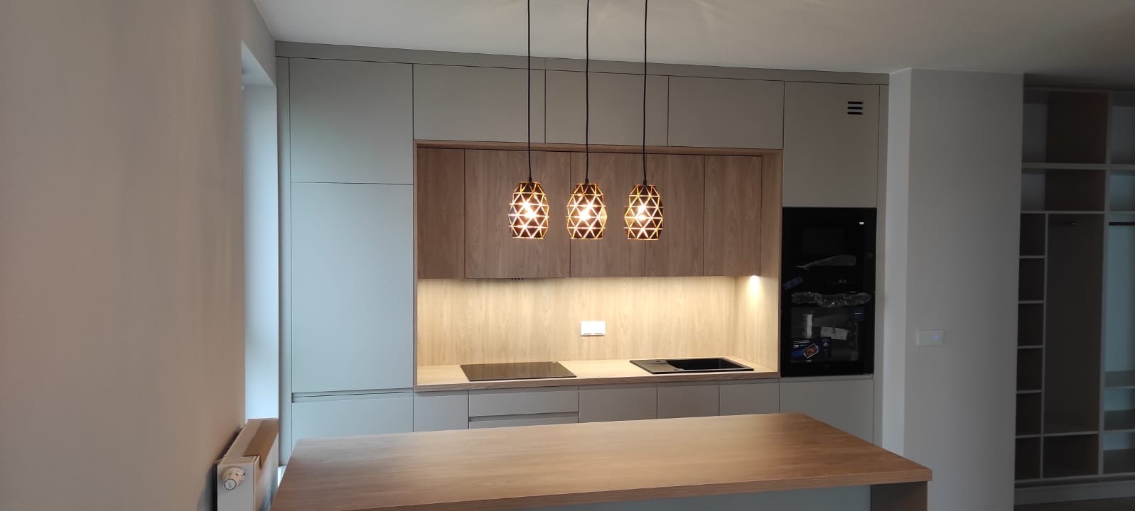 Nowoczesna kuchnia z drewnianymi frontami i trzema designerskimi lampami. Jasne szafki, płyta indukcyjna i piekarnik w zabudowie. Widoczna wyspa kuchenna.