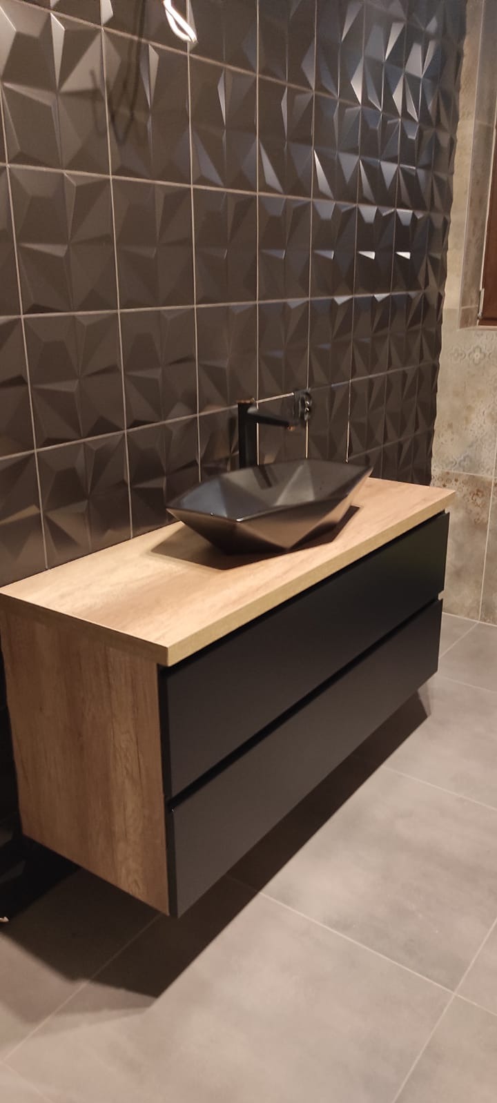 Nowoczesna szafka łazienkowa z umywalką w kształcie rombu i czarną armaturą na tle ściany z geometrycznych płytek. Fronty szafki w kolorze czarnym, blat w odcieniu drewna.
