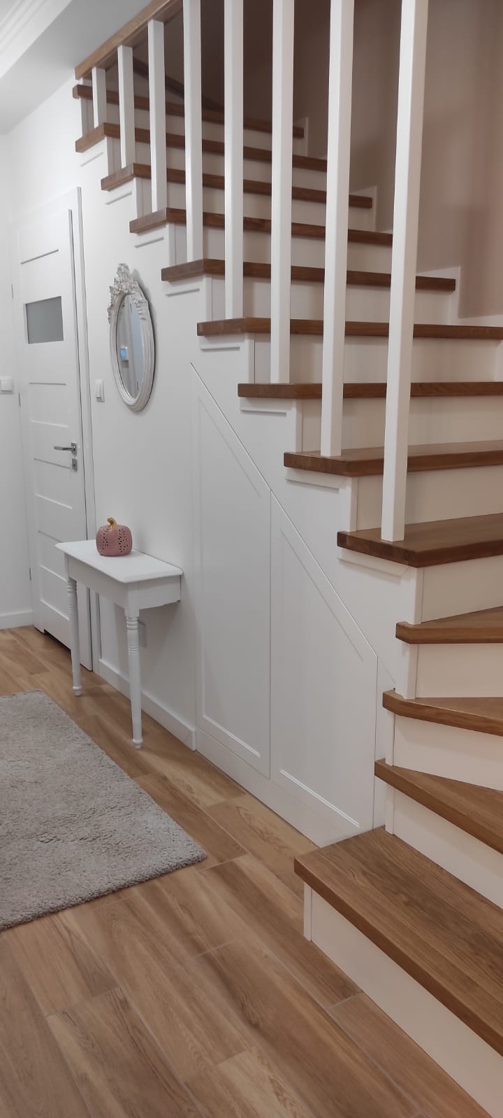 Drewniane schody z białymi balustradami i zabudową pod schodami. Dekoracyjny stolik z lustrem i ceramiczną dynią. Jasne wnętrze, drewniana podłoga i białe ściany.