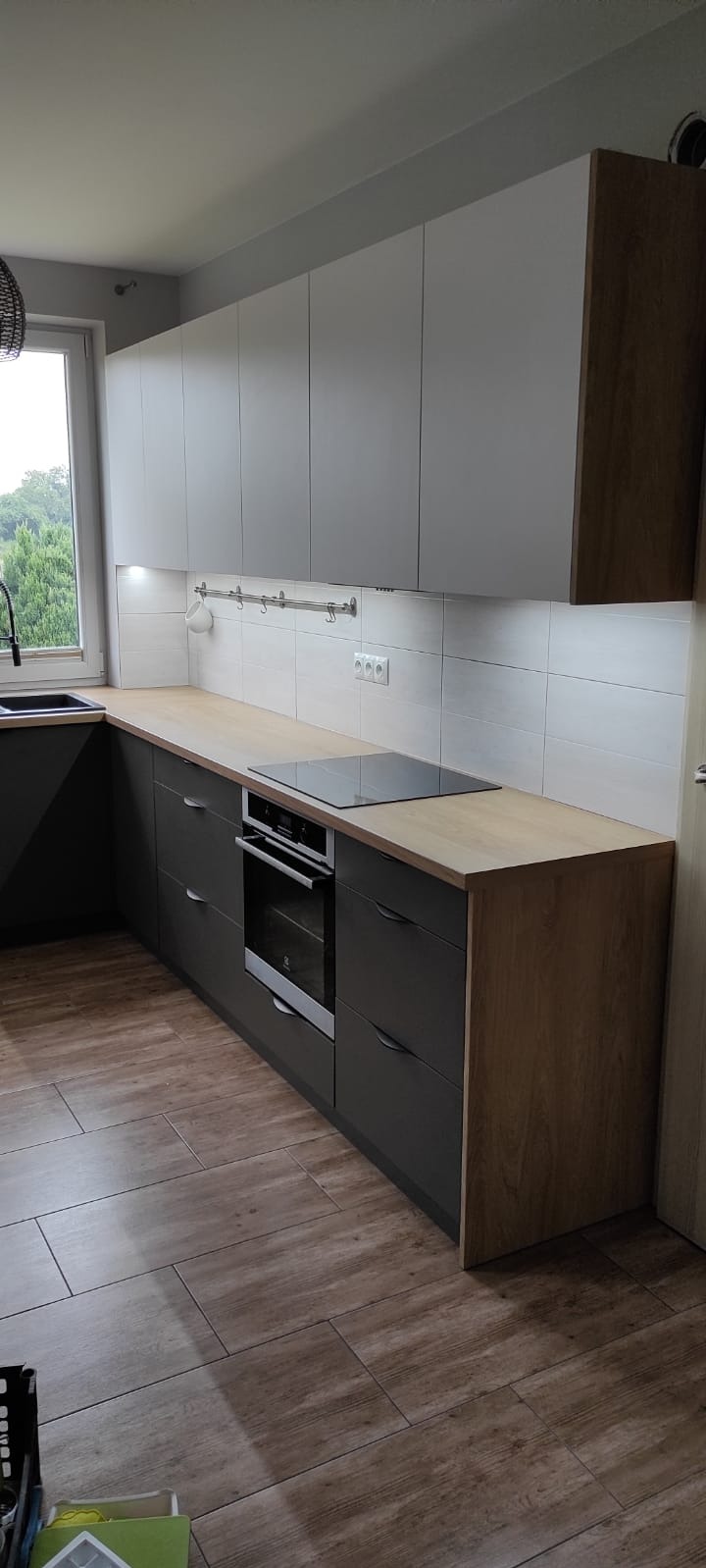 Nowoczesna kuchnia z szarymi szafkami i drewnianym blatem, płyta indukcyjna i piekarnik w zabudowie, jasne płytki na ścianie i drewnopodobna podłoga.