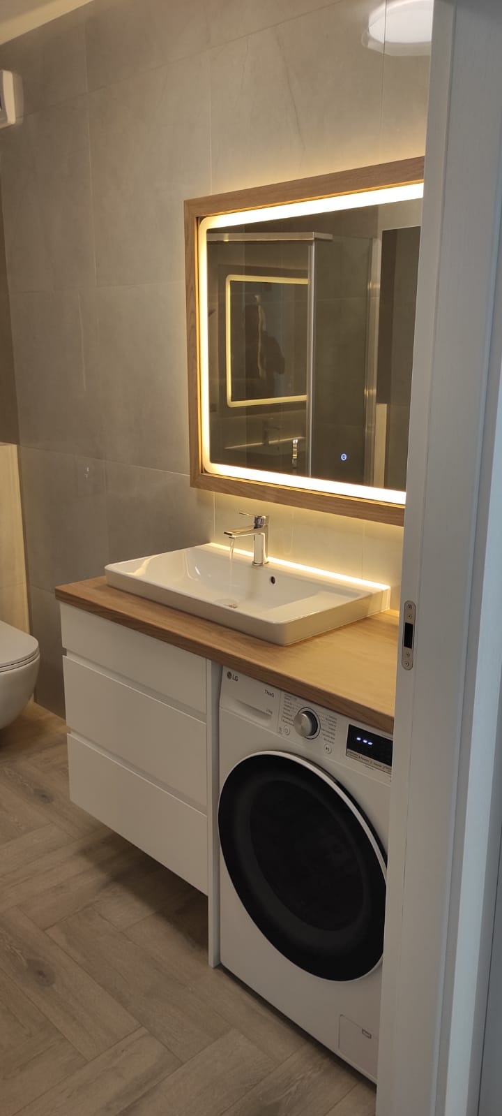 Nowoczesna łazienka z umywalką nablatową na szafce z pralką, podświetlane lustro w drewnianej ramie. Szare płytki i podłoga imitująca drewno.