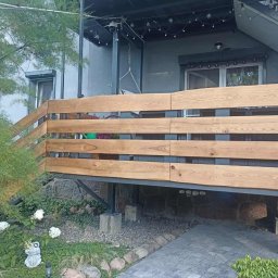 PA-TERA - Drewniana balustrada tarasu z poziomych desek o naturalnym odcieniu, wsparta na metalowej konstrukcji. Widok na ogród z roślinami i kamienną ścieżką.