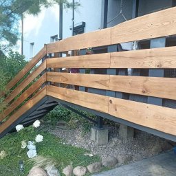 PA-TERA - Drewniana balustrada schodowa z metalową konstrukcją, widoczna z poziomu ogrodu. Jasne drewno kontrastuje z szarym metalem. W tle zieleń i fragment budynku.