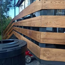 PA-TERA - Drewniana balustrada z poziomymi deskami, mocowana na metalowych słupkach, widoczna z boku, z elementami otoczenia w tle. Deski o wyraźnym usłojeniu.