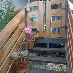 PA-TERA - Drewniane schody z balustradą i furtką, zdobione kwiatami w koszach. Widoczne metalowe elementy konstrukcyjne i naturalne oświetlenie.