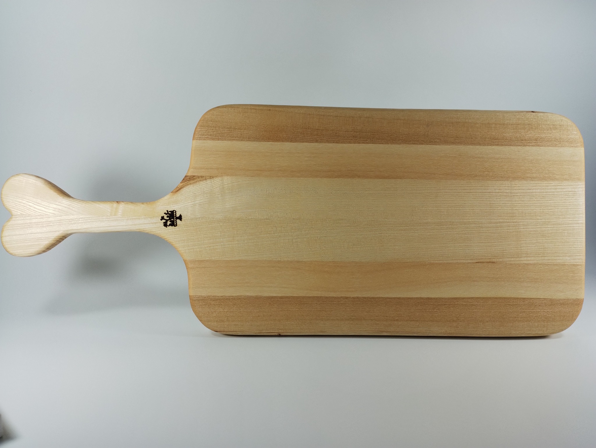 Drewniana deska do krojenia z uchwytem w kształcie kości, wykonana z jasnego drewna z widocznym usłojeniem. Na desce widoczne logo producenta. Minimalistyczne tło.
