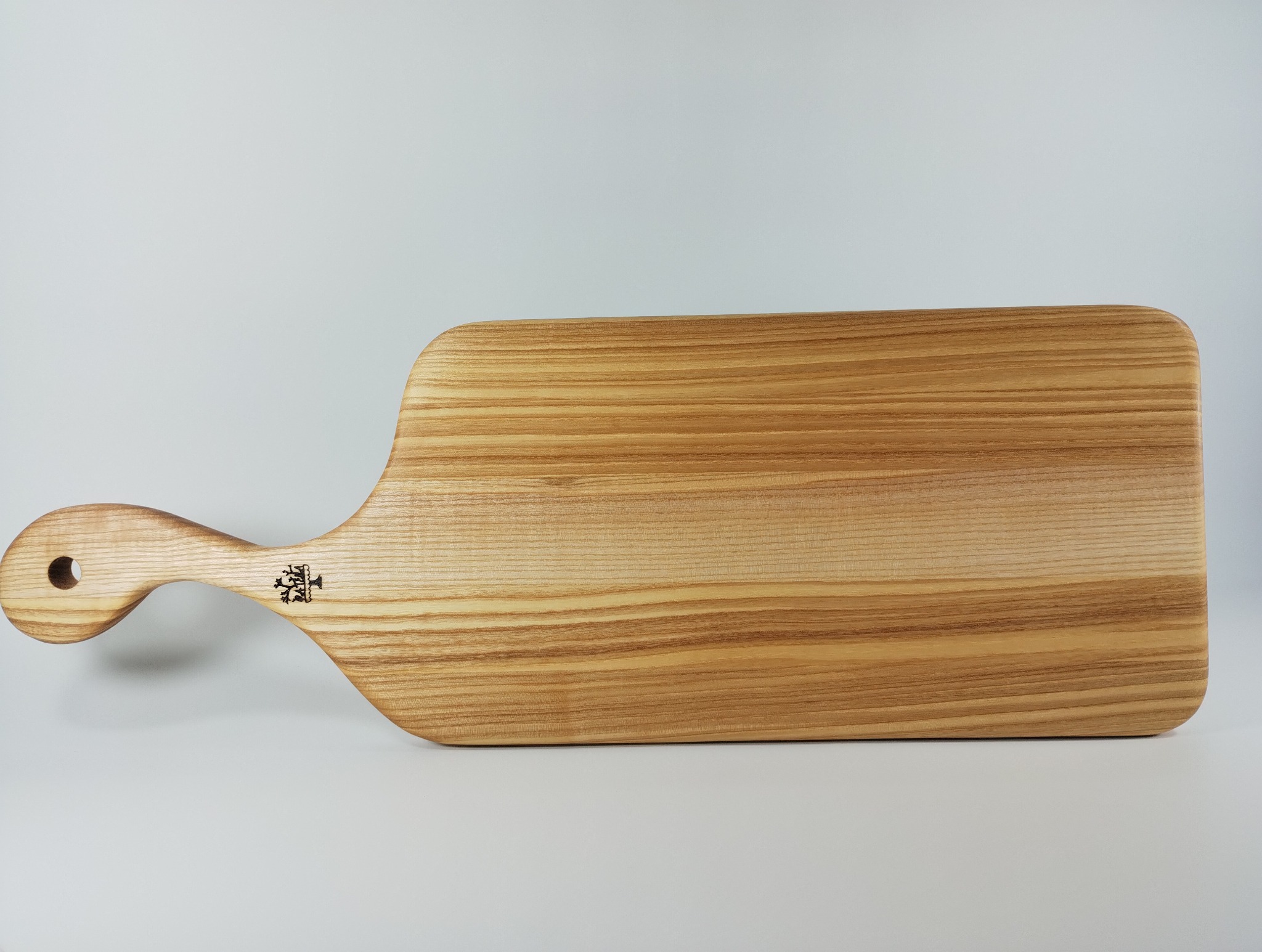 Drewniana deska do krojenia z uchwytem i wygrawerowanym logo, leżąca poziomo na jasnym tle. Widoczne naturalne usłojenie drewna.