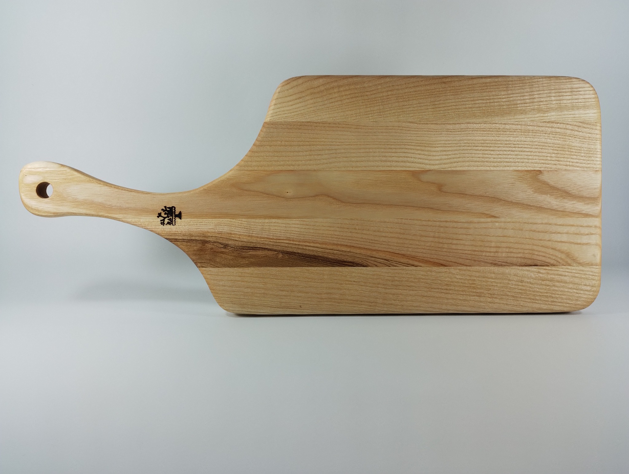 Drewniana deska do krojenia z uchwytem i wygrawerowanym logo, leżąca na jasnym tle. Widoczna struktura drewna i otwór w uchwycie.