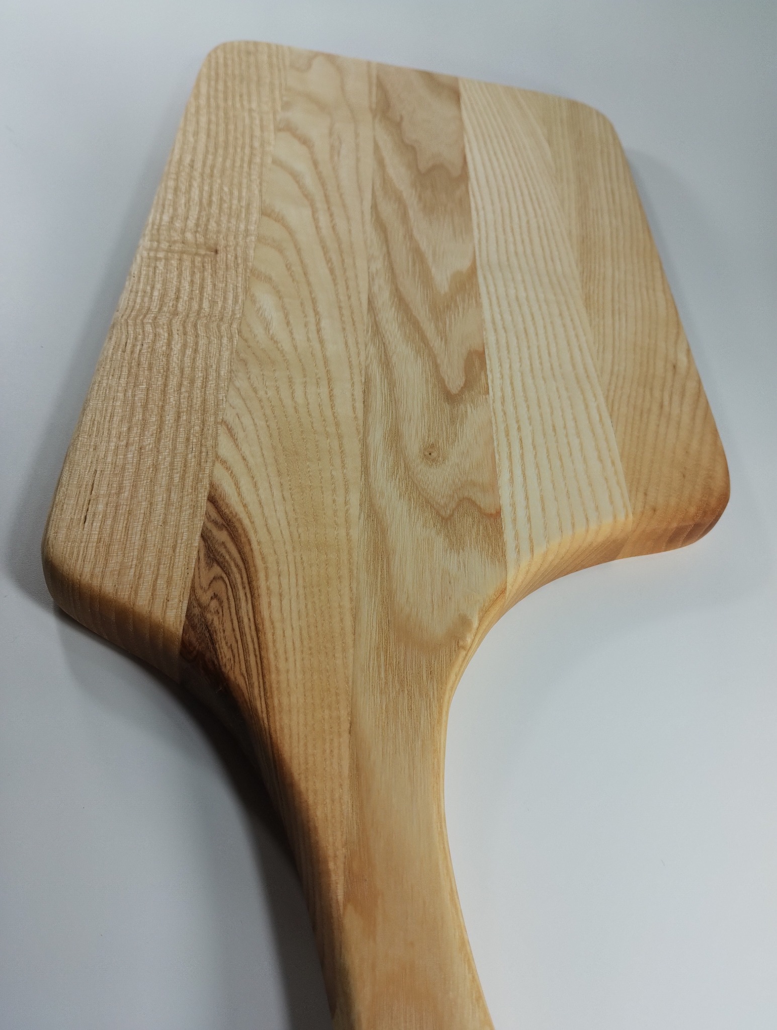 Jasna, drewniana deska do serwowania z uchwytem, ukazująca naturalny rysunek drewna. Unikalny kształt deski z zaokrąglonymi krawędziami. Widoczne detale obróbki drewna.