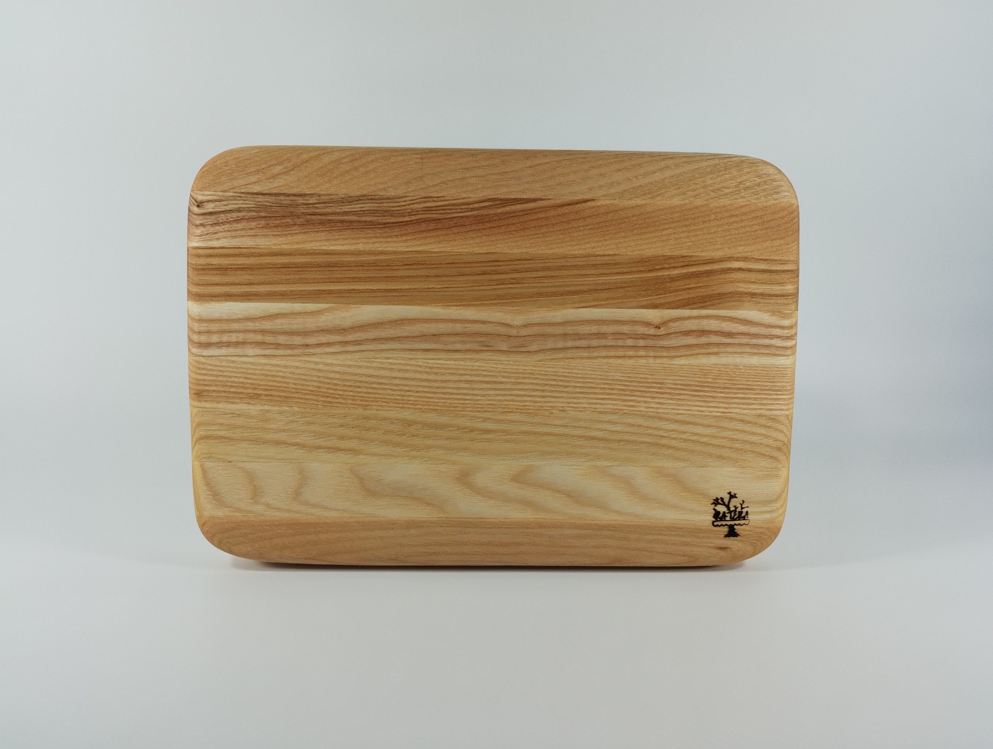 Drewniana deska do krojenia z zaokrąglonymi rogami i delikatnym logo w prawym dolnym rogu, prezentowana na jasnym tle. Widoczna struktura drewna.