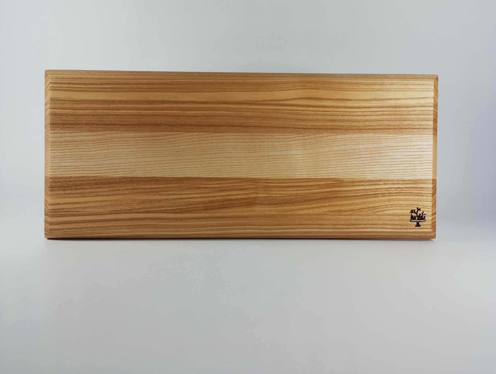 Prostokątny blat z jasnego drewna jesionowego z wyraźnym usłojeniem, wykończony na gładko, z delikatnym logo w prawym dolnym rogu, na jasnym tle.
