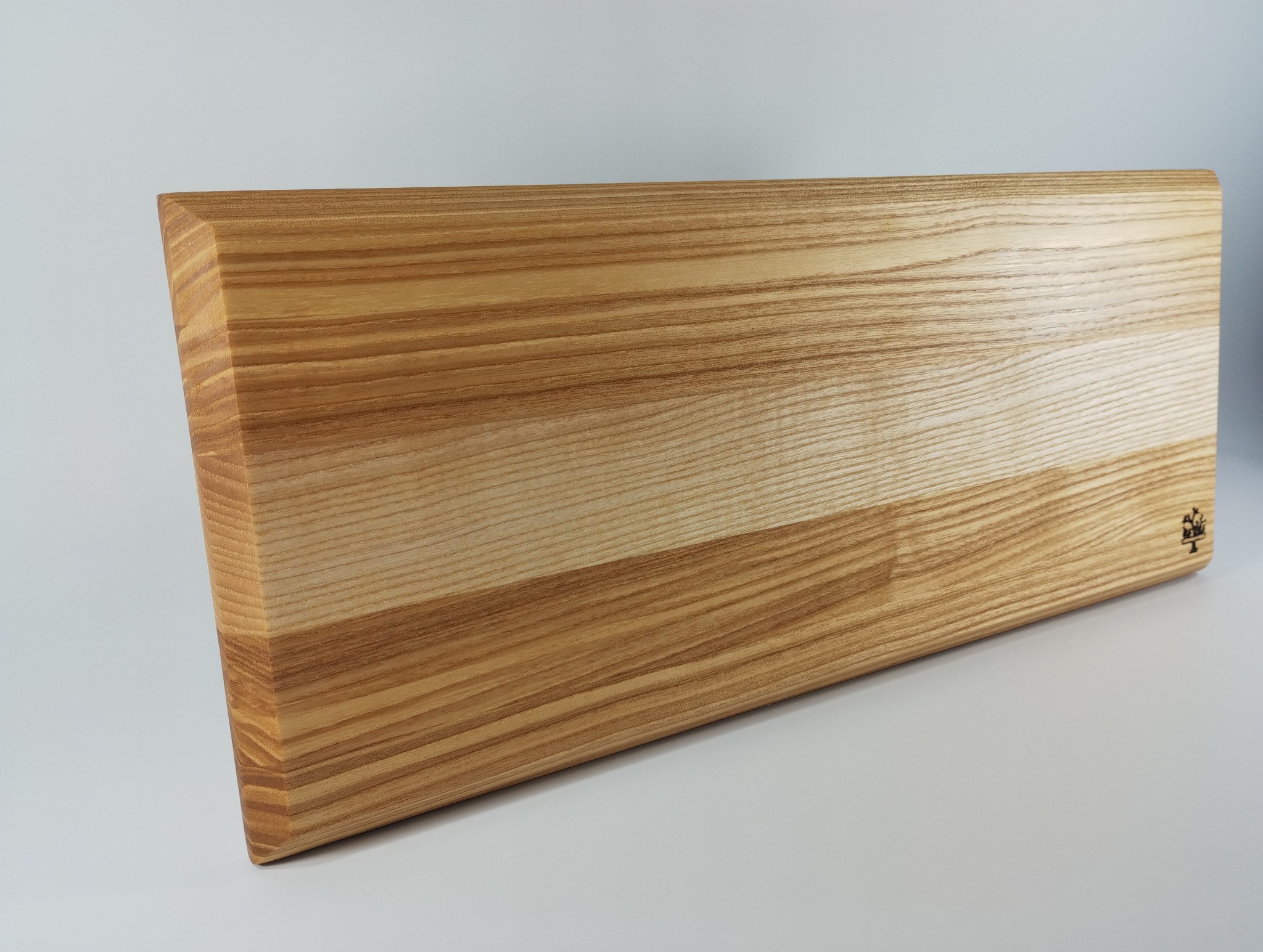 Drewniany blat z wyraźnym usłojeniem, wykończony na gładko, z zaokrąglonymi krawędziami i delikatnym logo w prawym dolnym rogu, prezentowany na jasnym tle.