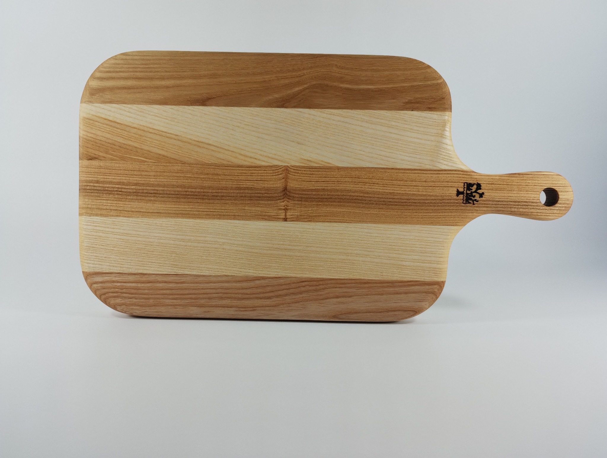 Drewniana deska do krojenia z uchwytem, wykonana z jasnego drewna z ciemniejszymi paskami. Na uchwycie widoczne logo. Deska leży na białym tle.