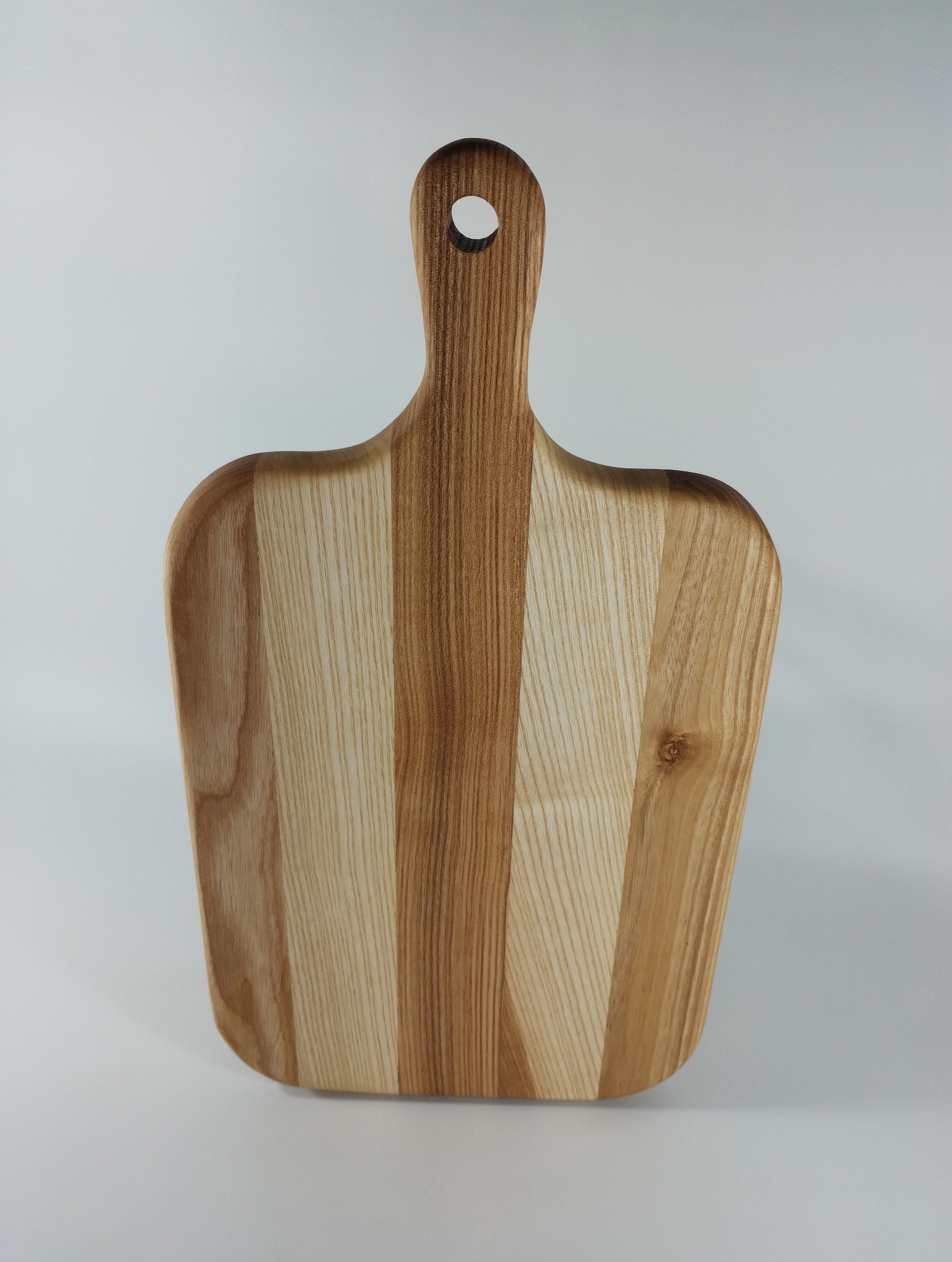 Drewniana deska do krojenia z uchwytem i otworem, wykonana z jasnego drewna o widocznych słojach, na jednolitym, jasnym tle. Minimalistyczny design.