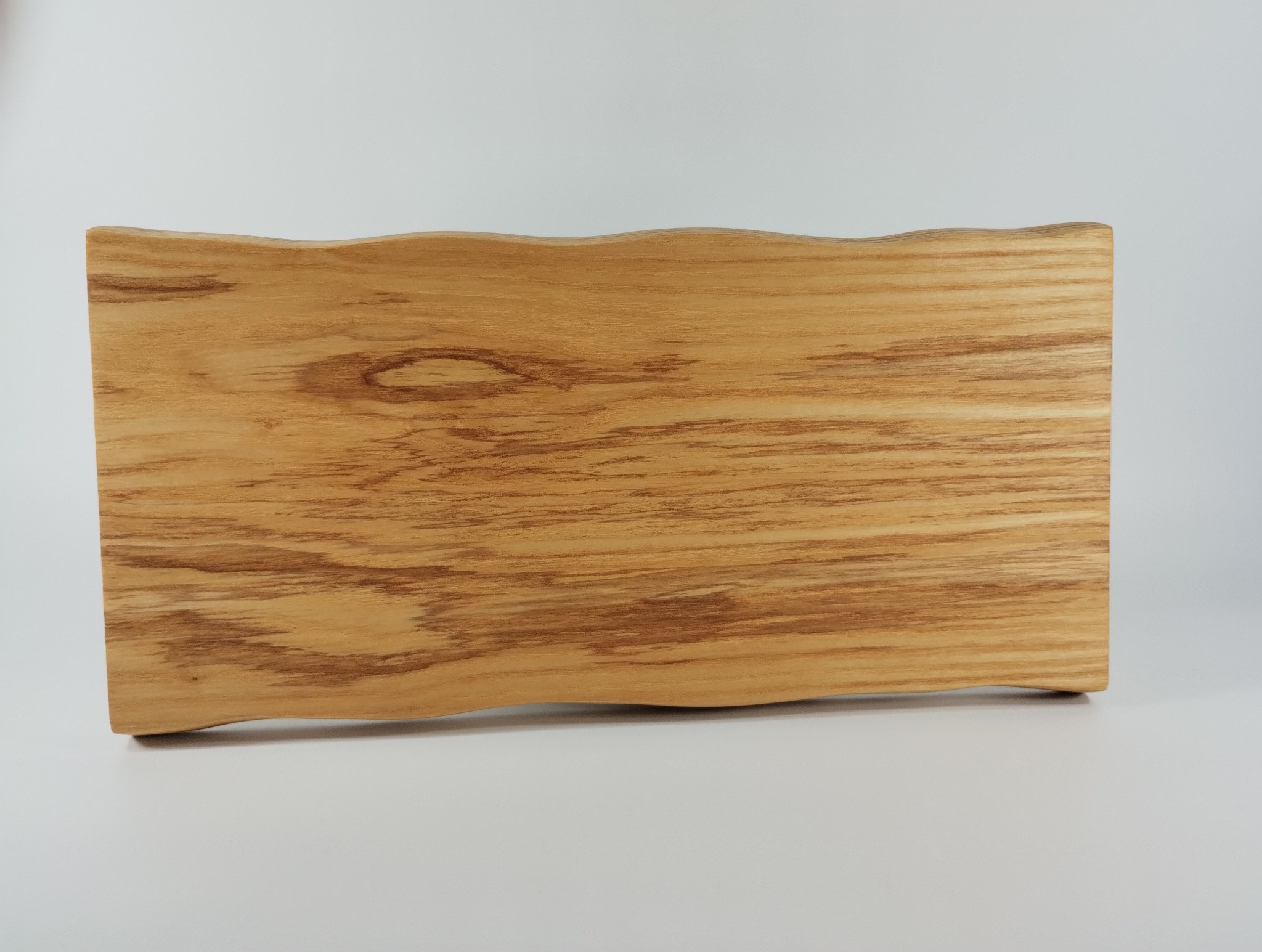 Drewniany blat o nieregularnym, falistym kształcie krawędzi, ukazujący naturalny rysunek słojów. Jasne drewno na neutralnym tle.
