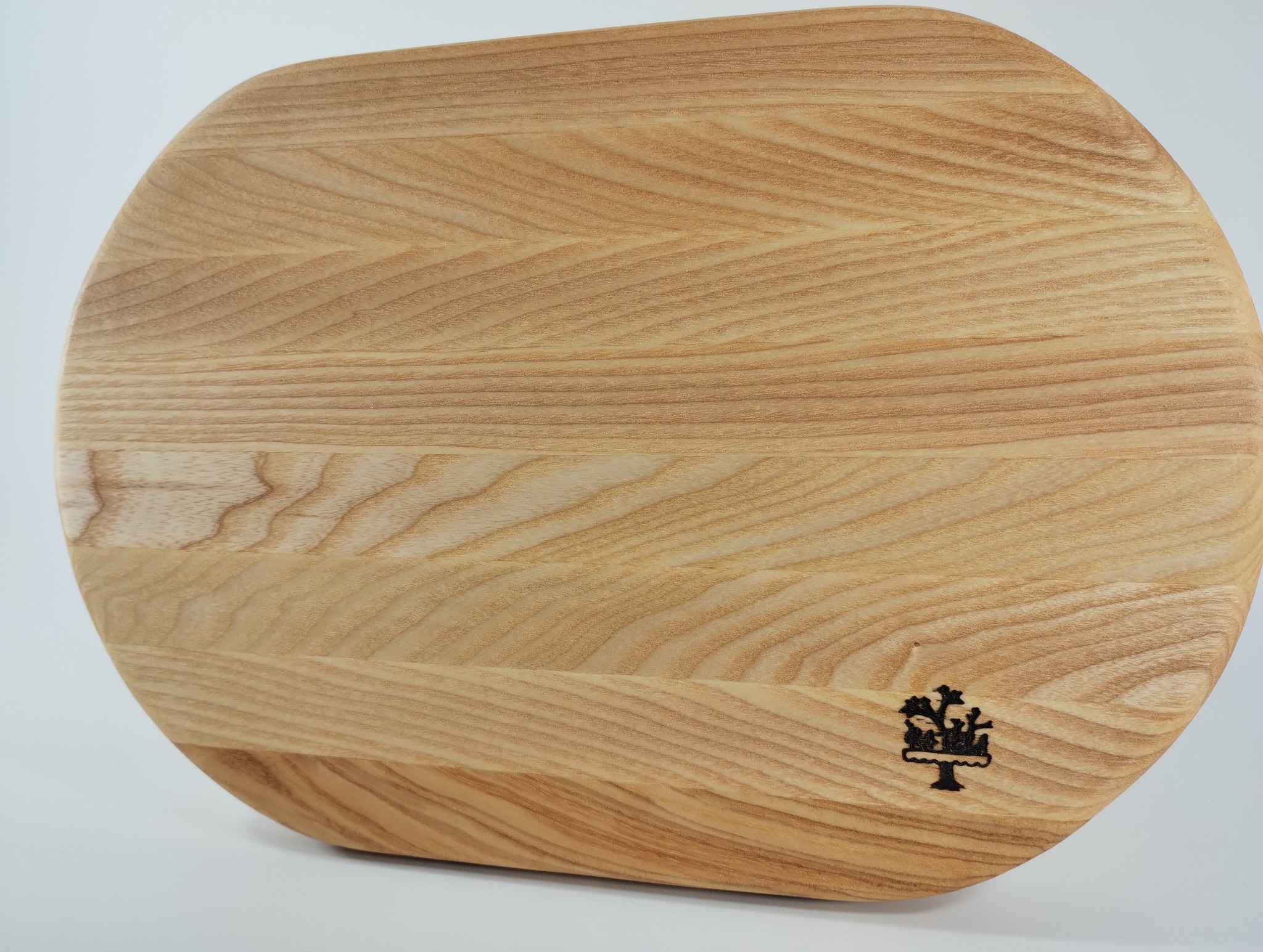 Jasny, okrągły blat z drewna jesionowego z zaokrąglonymi krawędziami i wypalonym logo w prawym dolnym rogu, ukazujący unikalny wzór słojów.