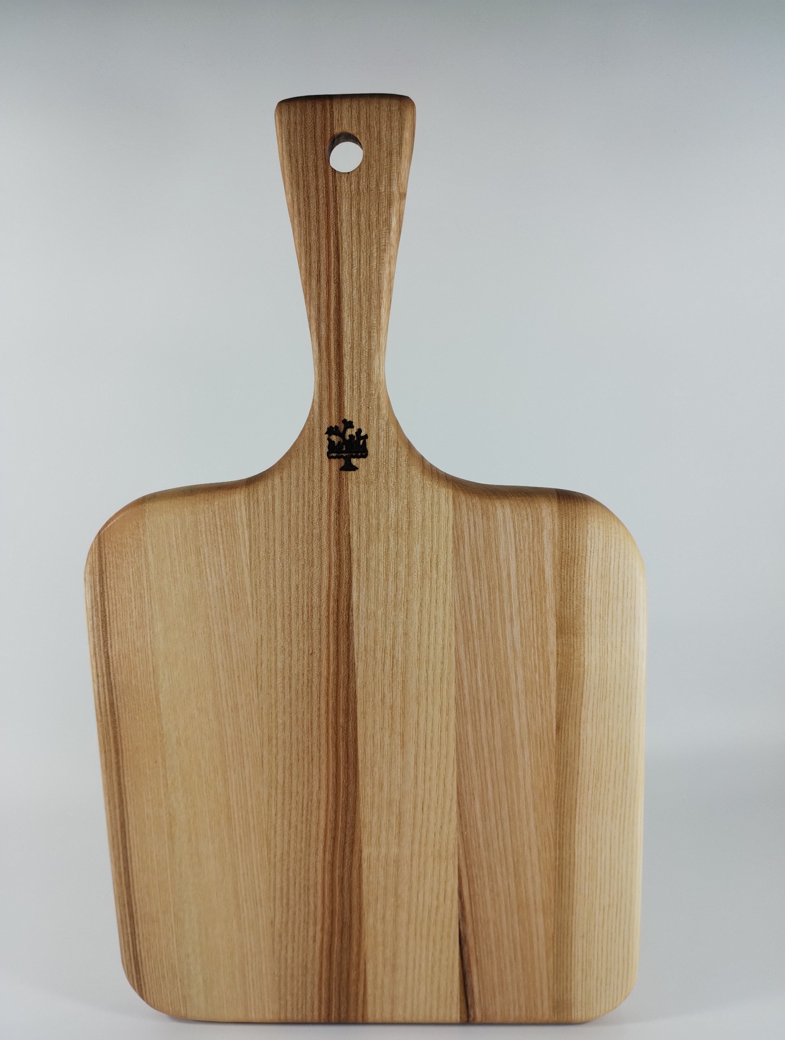 Drewniana deska do krojenia z uchwytem i otworem do zawieszenia, z delikatnym logo w centralnej części. Jasne drewno z widocznym usłojeniem na neutralnym tle.