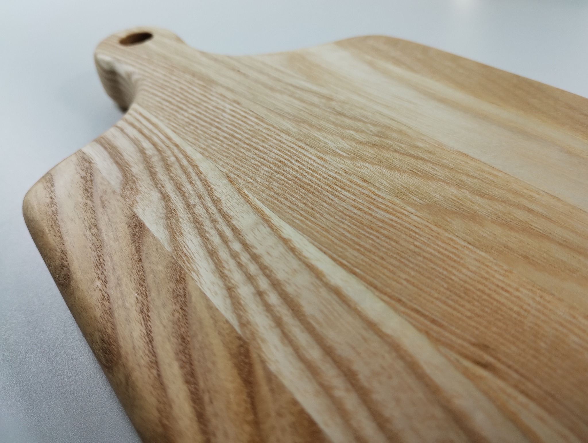 Jasna, drewniana deska do krojenia z uchwytem i otworem, ukazująca naturalny rysunek słojów drewna, sfotografowana pod kątem na gładkim, jasnym tle.