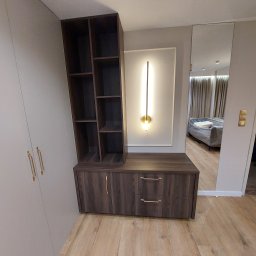 Custom-Made Furniture Meblowo - Meble na wymiar - Ciemna szafka z półkami i szufladami, złote uchwyty, oświetlenie dekoracyjne na ścianie, lustro odbijające wnętrze pokoju z sofą pod folią ochronną.