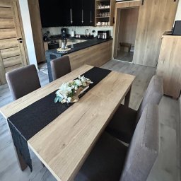 Custom-Made Furniture Meblowo - Meble na wymiar - Nowoczesna kuchnia z drewnianymi frontami i czarnymi akcentami, widok na stół z dekoracją kwiatową i krzesła. Przesuwne drzwi w tle.