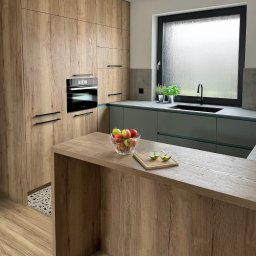 Custom-Made Furniture Meblowo - Meble na wymiar - Nowoczesna kuchnia z drewnianymi frontami, czarnymi uchwytami i oknem z widokiem na zewnątrz. Na blacie szklana misa z jabłkami i deska z przekrojonym jabłkiem.