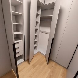 Jasnoszara szafa narożna z otwartymi drzwiami, widoczne półki i szuflady z czarnymi uchwytami, na podłodze panele w odcieniach drewna.