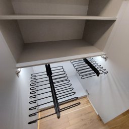 Wnętrze szafy z wysuwanymi wieszakami na spodnie, zamontowanymi pod półką. Widoczne oświetlenie LED wewnątrz szafy i drewniana podłoga.