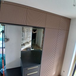 A&P Meble Matula Piotr - Nowoczesna zabudowa meblowa z jasnymi, pionowymi ryflowanymi panelami ściennymi, ciemnoszarą komodą ze złotymi uchwytami i wbudowanym lustrem, na tle parkietu w jodełkę.