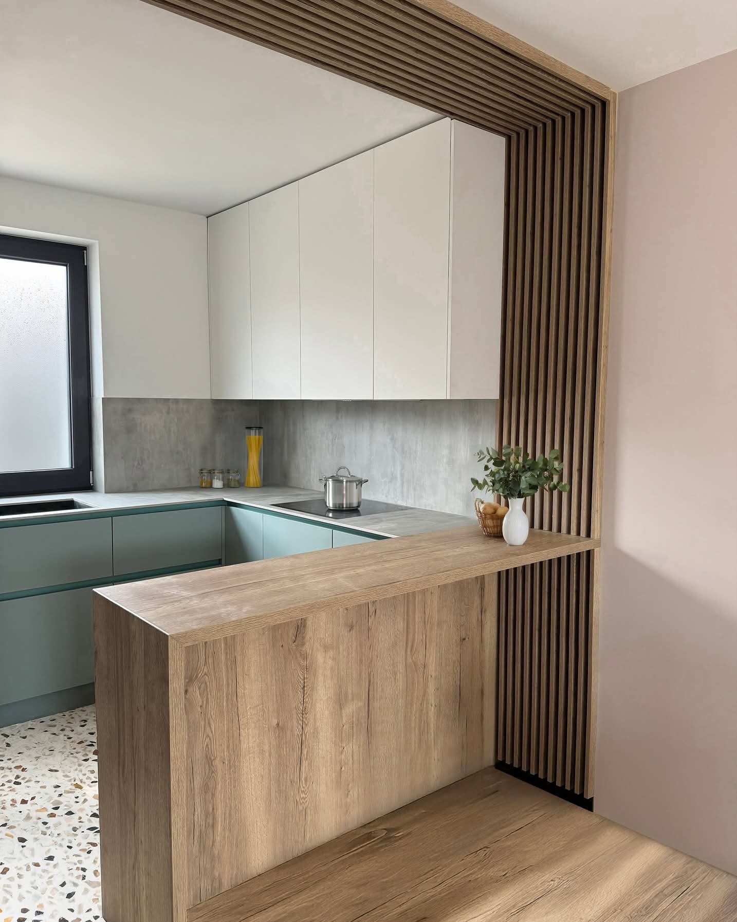 Nowoczesna kuchnia z zielonymi szafkami i drewnianym blatem oddzielającym przestrzeń, uzupełniona o drewniane lamele dekoracyjne.