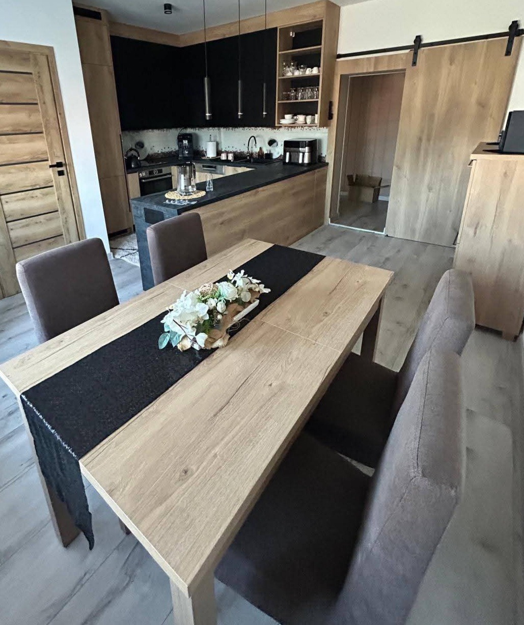 Nowoczesna kuchnia z drewnianymi szafkami, czarnym blatem i wyspą. Stół z dekoracją kwiatową na pierwszym planie. Przesuwne drzwi do spiżarni.