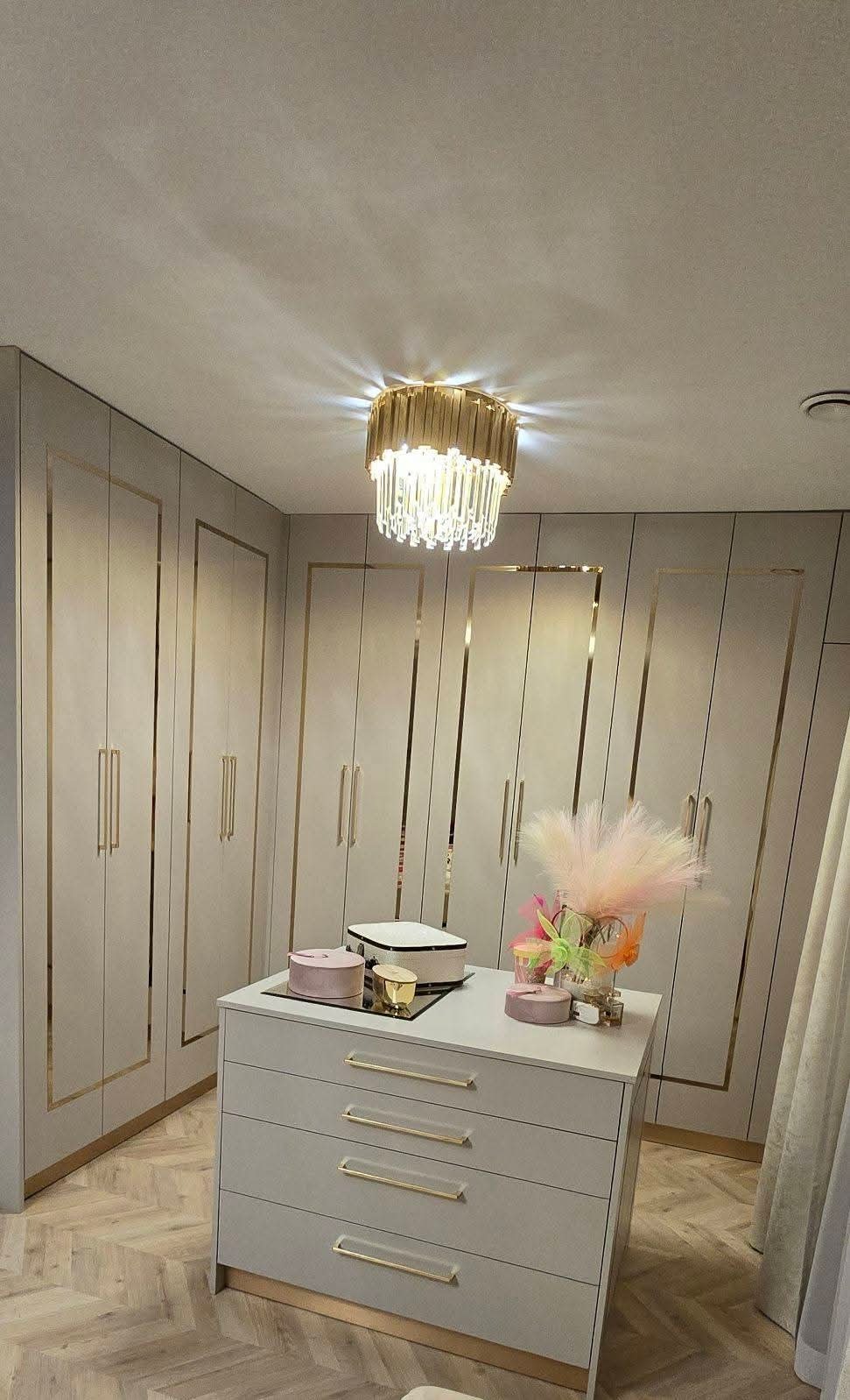 Elegancka garderoba z szarymi szafami wnękowymi, złotymi akcentami i kryształowym żyrandolem. Na komodzie dekoracje i pudełka.