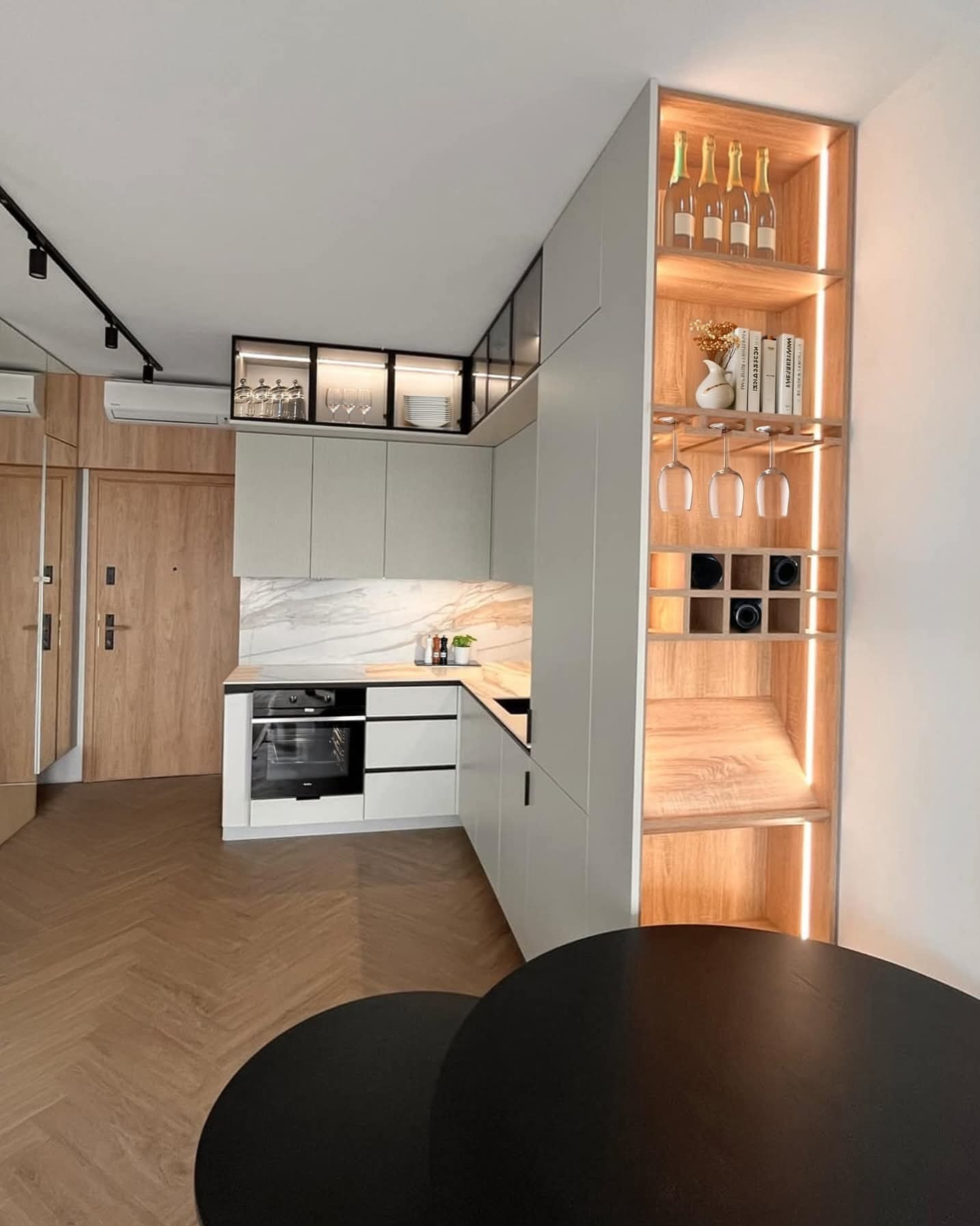 Nowoczesna kuchnia z szarymi szafkami, drewnianym regałem z oświetleniem LED i blatem imitującym marmur. Parkiet w jodełkę, czarny stół w tle.
