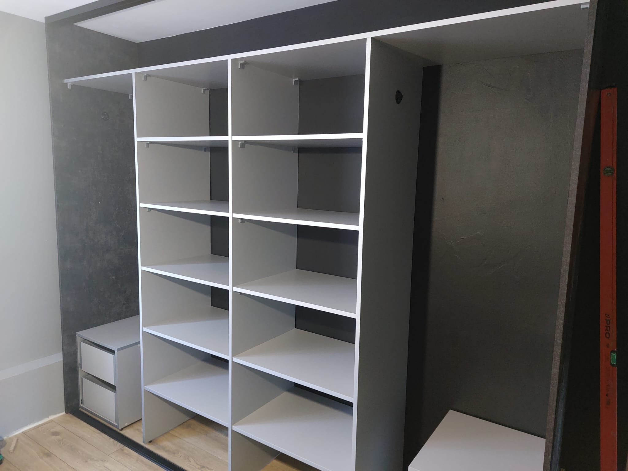 Wnętrze szafy wnękowej z półkami i szufladami, wykończonej w odcieniach szarości, z widoczną poziomicą po prawej stronie. Minimalistyczny design.