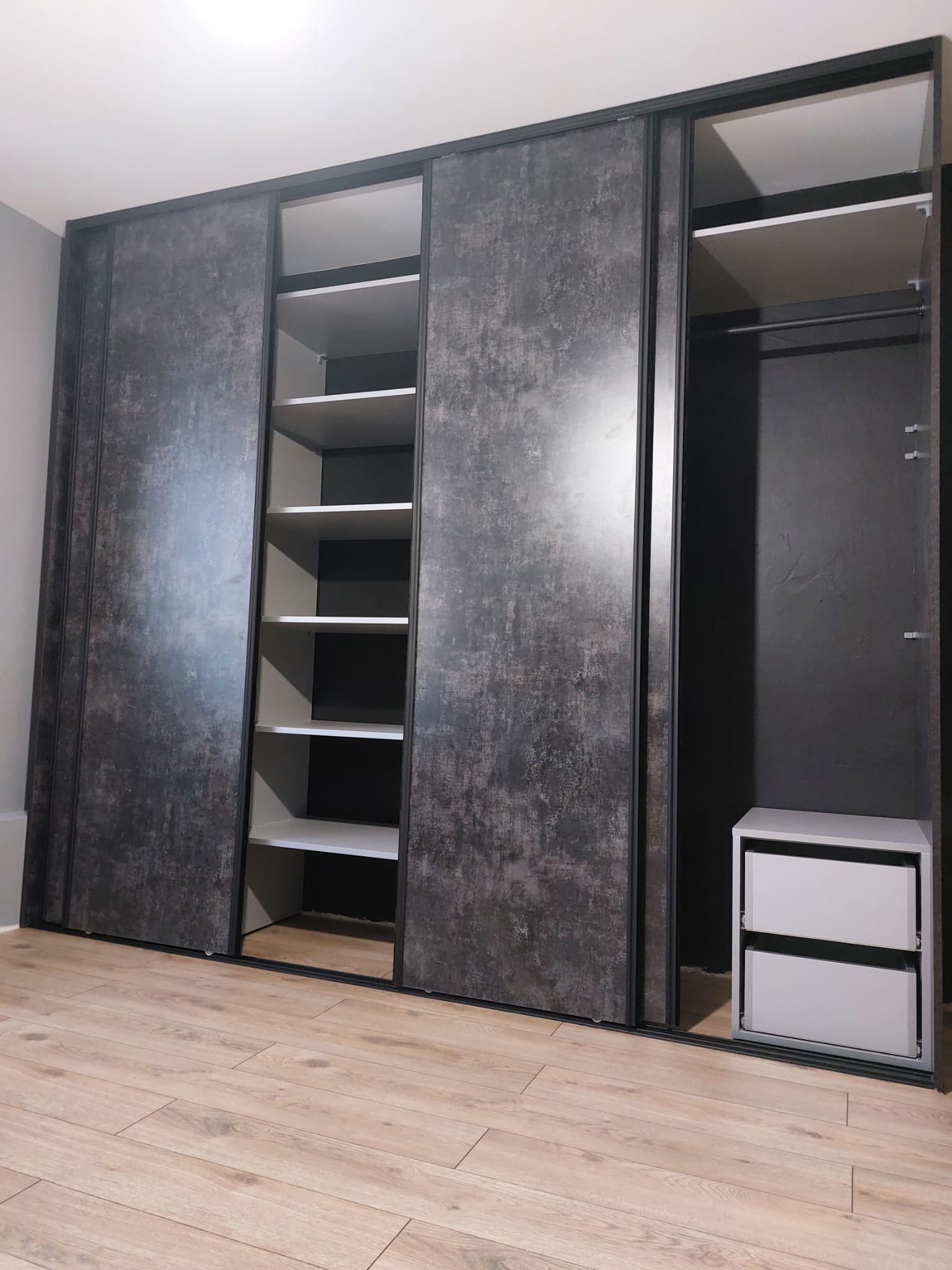 Nowoczesna szafa wnękowa z przesuwnymi drzwiami o fakturze betonu, z czarnymi obramowaniami, na podłodze z paneli. Widoczne półki i szuflady.