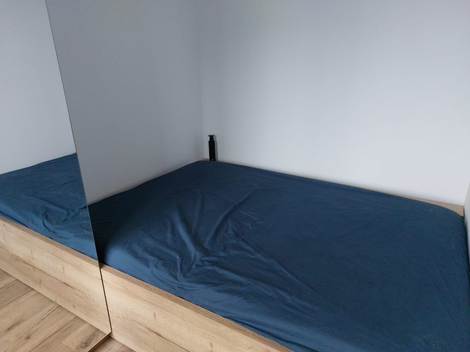 Łóżko z drewnianą ramą i niebieskim prześcieradłem, częściowo odbite w lustrze. Minimalistyczny design, jasne ściany, widoczny fragment podłogi.