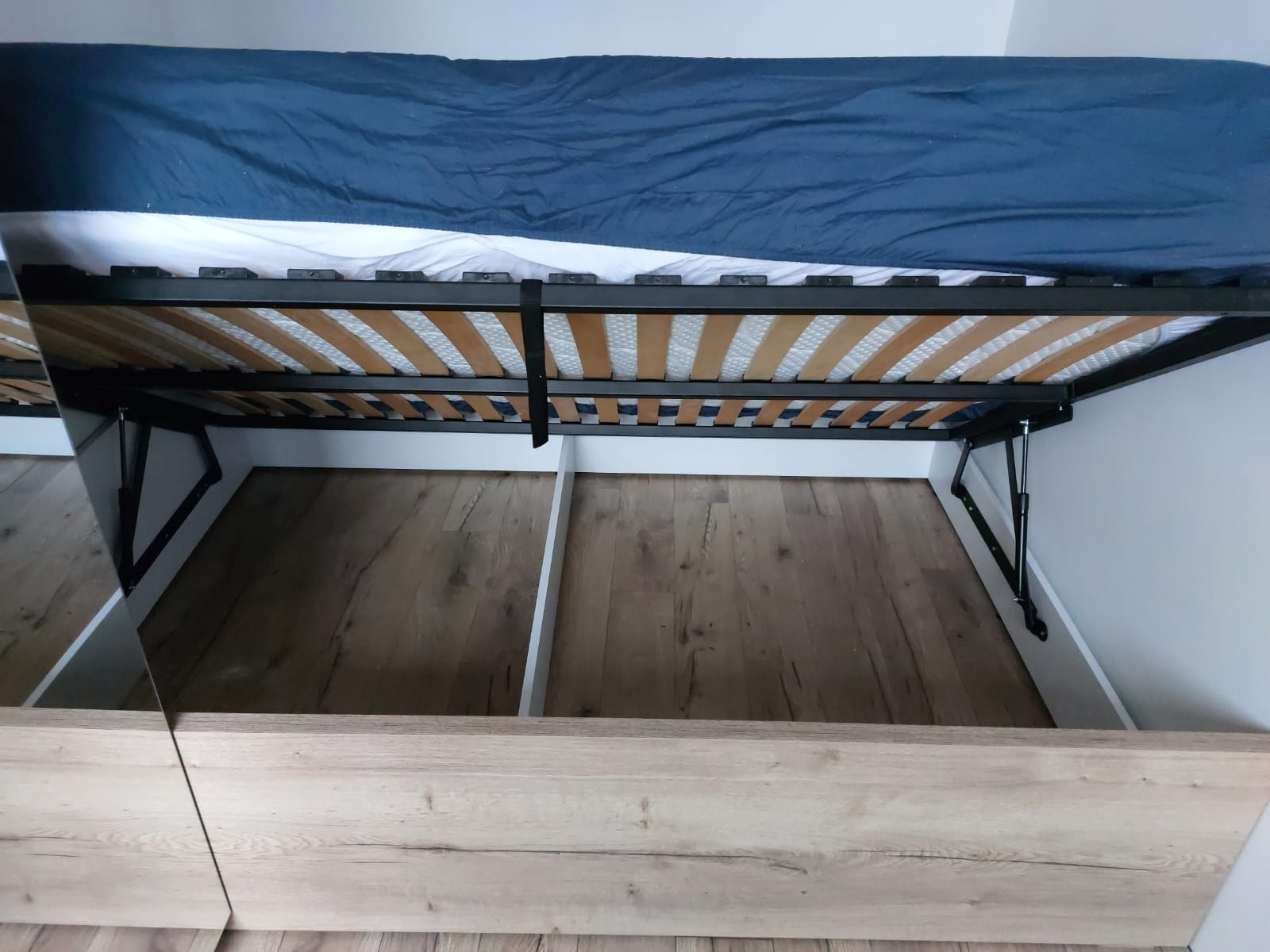 Otwarta rama łóżka z pojemnikiem, widoczne drewniane dno i mechanizm podnoszenia. Jasne drewno na froncie, wnętrze białe. Materac z niebieskim prześcieradłem na górze.