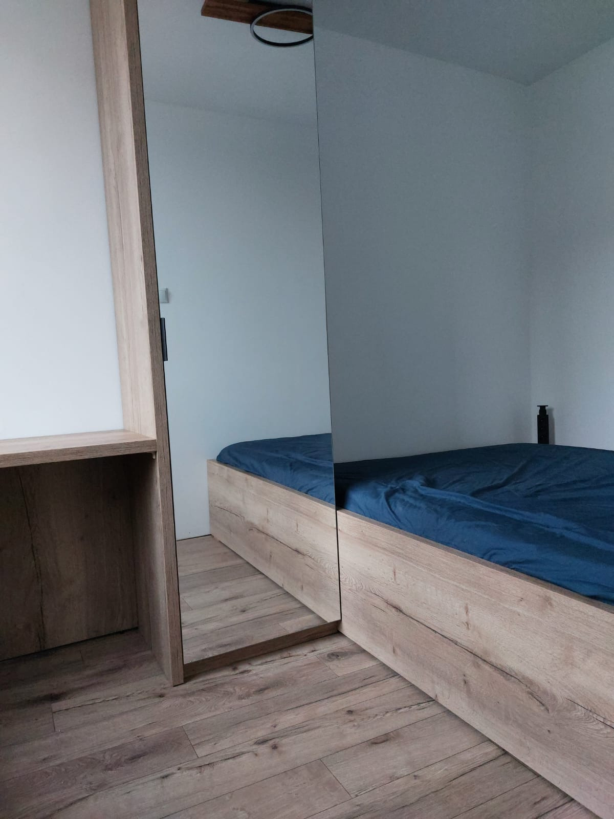 Szafa wnękowa z lustrzanymi drzwiami przesuwnymi, odbijająca łóżko z drewnianą ramą i niebieską pościelą. Widoczny fragment biurka z półkami i podłoga z paneli.
