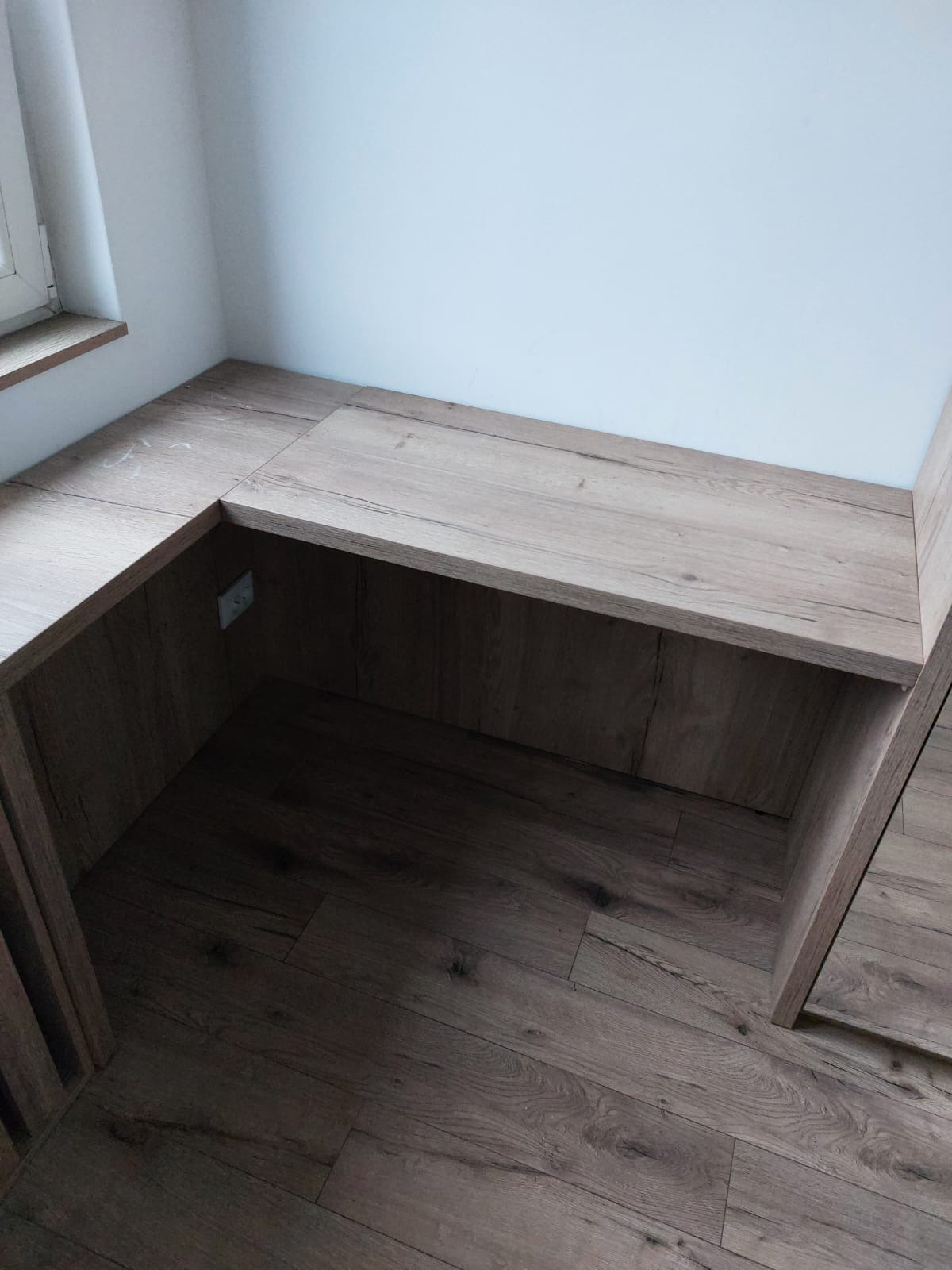 Drewniana zabudowa wnęki z blatem w kształcie litery L, dopasowana do podłogi z paneli. Widoczne gniazdko elektryczne. Ujęcie z góry, podkreślające detale wykończenia.