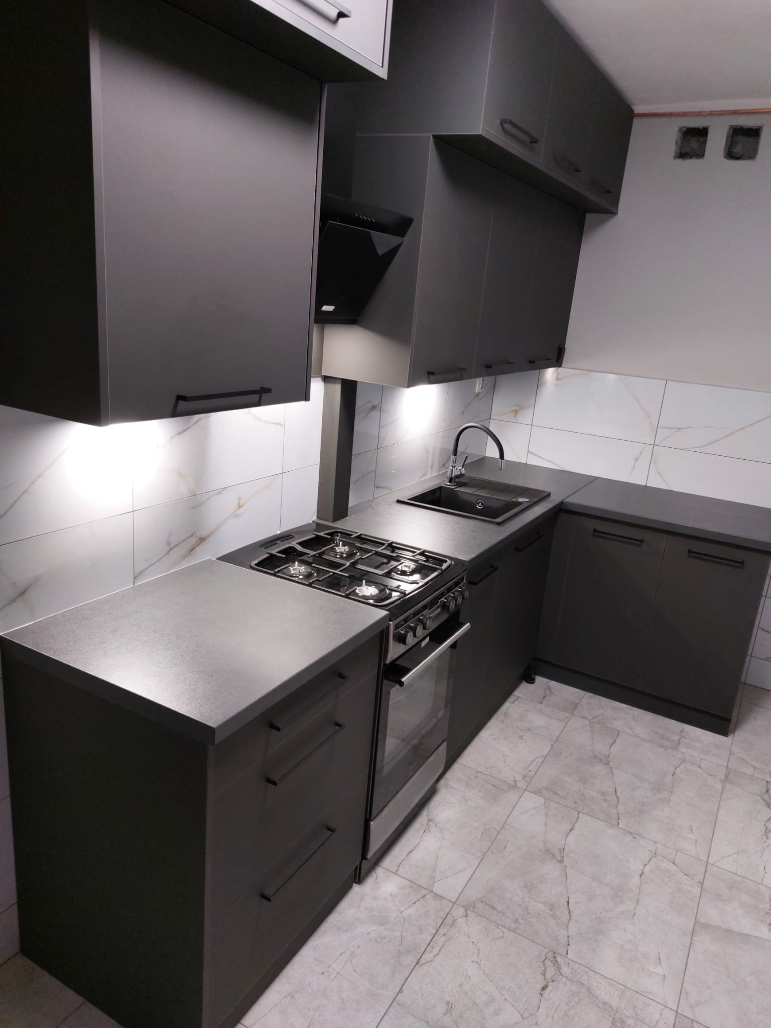 Grafitowa kuchnia na wymiar z marmurowymi płytkami. Ujęcie z perspektywy, prezentujące zabudowę kuchenną z piekarnikiem i płytą gazową.