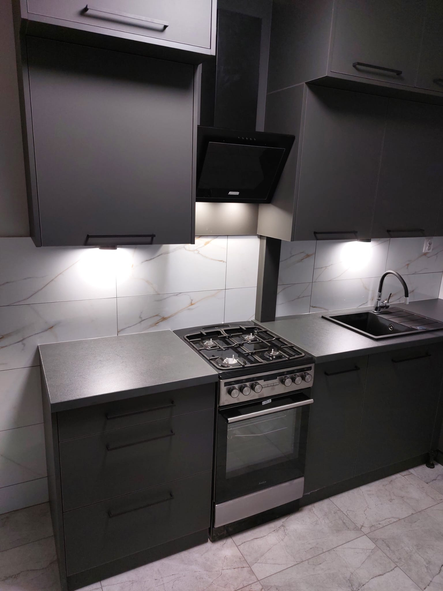 Nowoczesna, szara kuchnia z marmurowymi płytkami nad blatem. Uwagę przyciągają minimalistyczne uchwyty szafek i czarny okap nad płytą gazową.