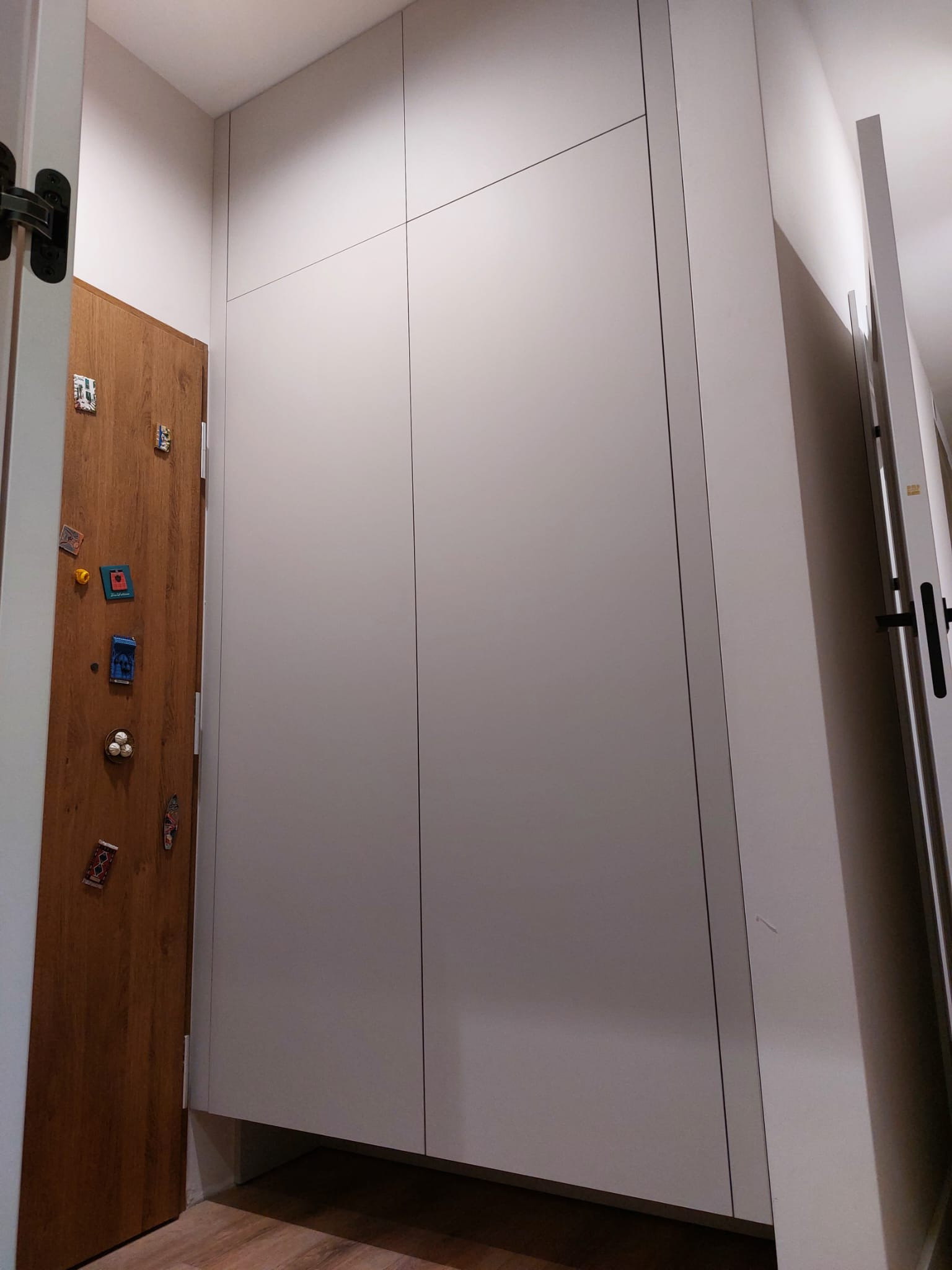 Biała szafa wnękowa z minimalistycznymi frontami, sąsiadująca z drewnianymi drzwiami ozdobionymi magnesami, tworząca spójną aranżację przedpokoju.
